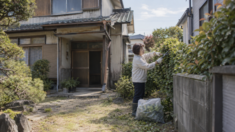 草木が伸びた空き家の外観と、見回りに訪れた所有者