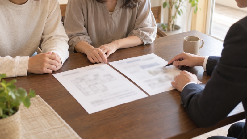 住宅ローンの借り方を相談する30代の夫婦と担当者。こげ茶の木のテーブルに2通の契約書類が並ぶ打合せの様子