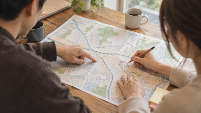 住宅街の地図の上で駅からの道を指でたどる夫婦の手元