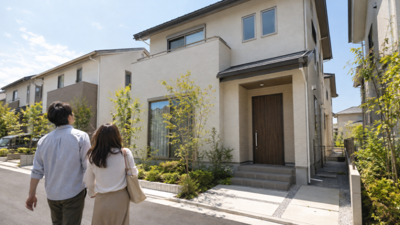 分譲地の新築住宅の前の道を、夫婦が建物を見上げながら歩いている後ろ姿（イメージ）