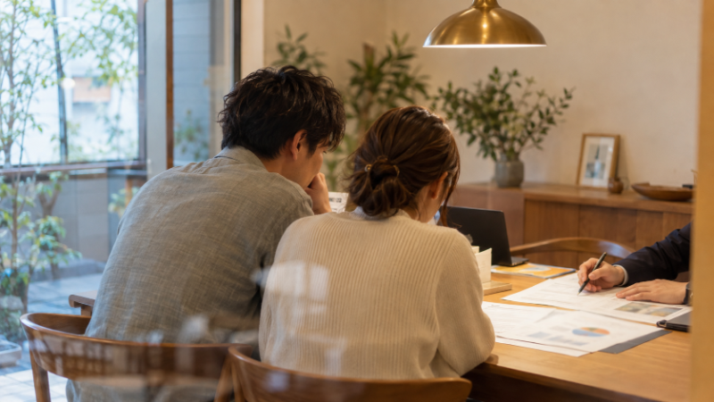 打合せスペースで、夫婦と担当者が資金計画の書類をのぞき込んでいる様子（イメージ）