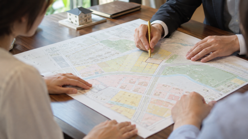 打合せの机で、担当者の手が都市計画図の上に敷地の形を描き、施主がのぞき込んでいる手元（イメージ）
