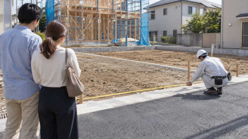分譲地の区画の前で、担当者が巻尺を伸ばして間口を測り、夫婦が少し離れて見ている様子（イメージ）